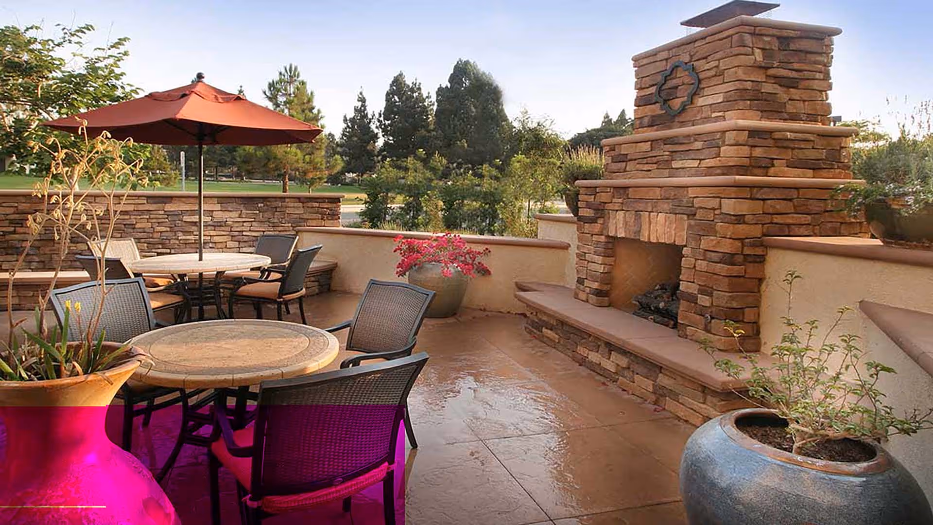 Outdoor patio area with round tables and chairs, a large umbrella, potted plants, and a stone fireplace with a decorative emblem. Trees and greenery are visible in the background under a clear sky.