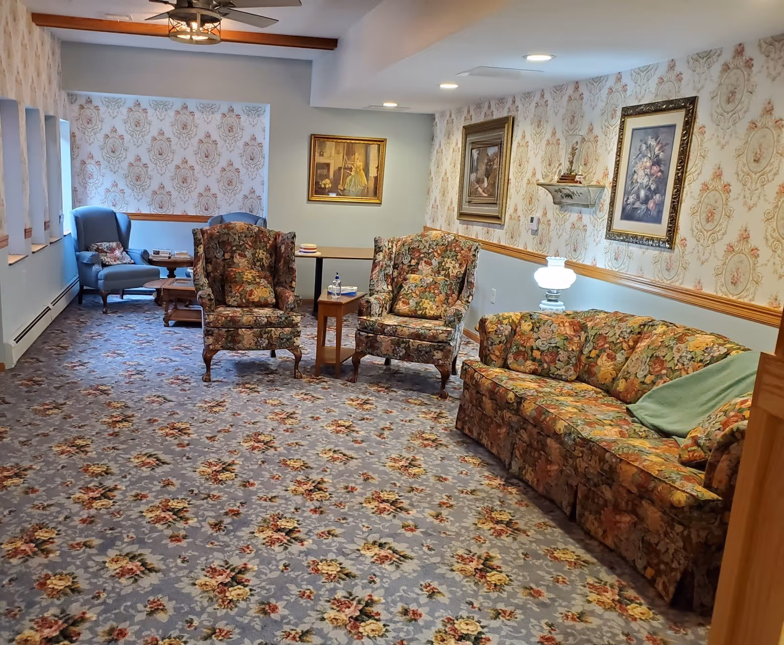 A cozy common living room with floral-patterned carpet, matching floral armchairs and sofa, framed paintings, and lamps.