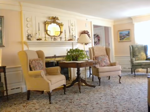 A cozy living room area with two beige upholstered armchairs and one green upholstered armchair arranged around a wooden table with a potted plant. The room features a fireplace with decorative items and a round mirror above it, a floor lamp, framed paintings on the walls, and a patterned carpet.