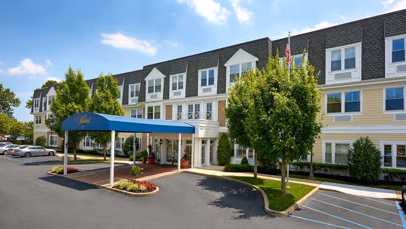Front entrance of a three-story assisted living building with a blue awning, parking lot, and trees.