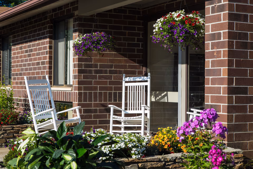 A brick building entrance with white rocking chairs on a porch surrounded by hanging baskets and colorful flowering plants.