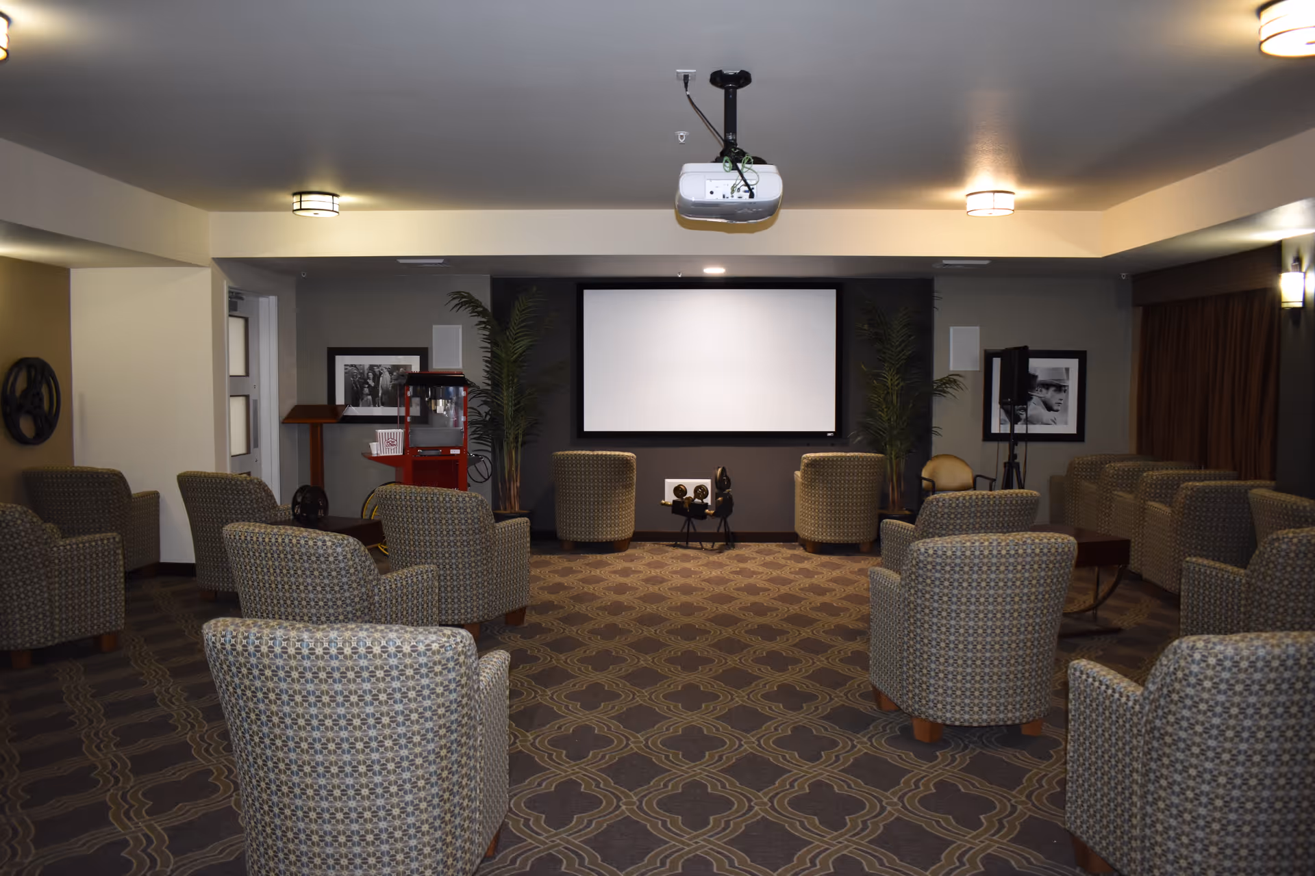 Small screening room with a ceiling projector, blank projection screen, a popcorn machine and multiple patterned armchairs arranged facing the screen.