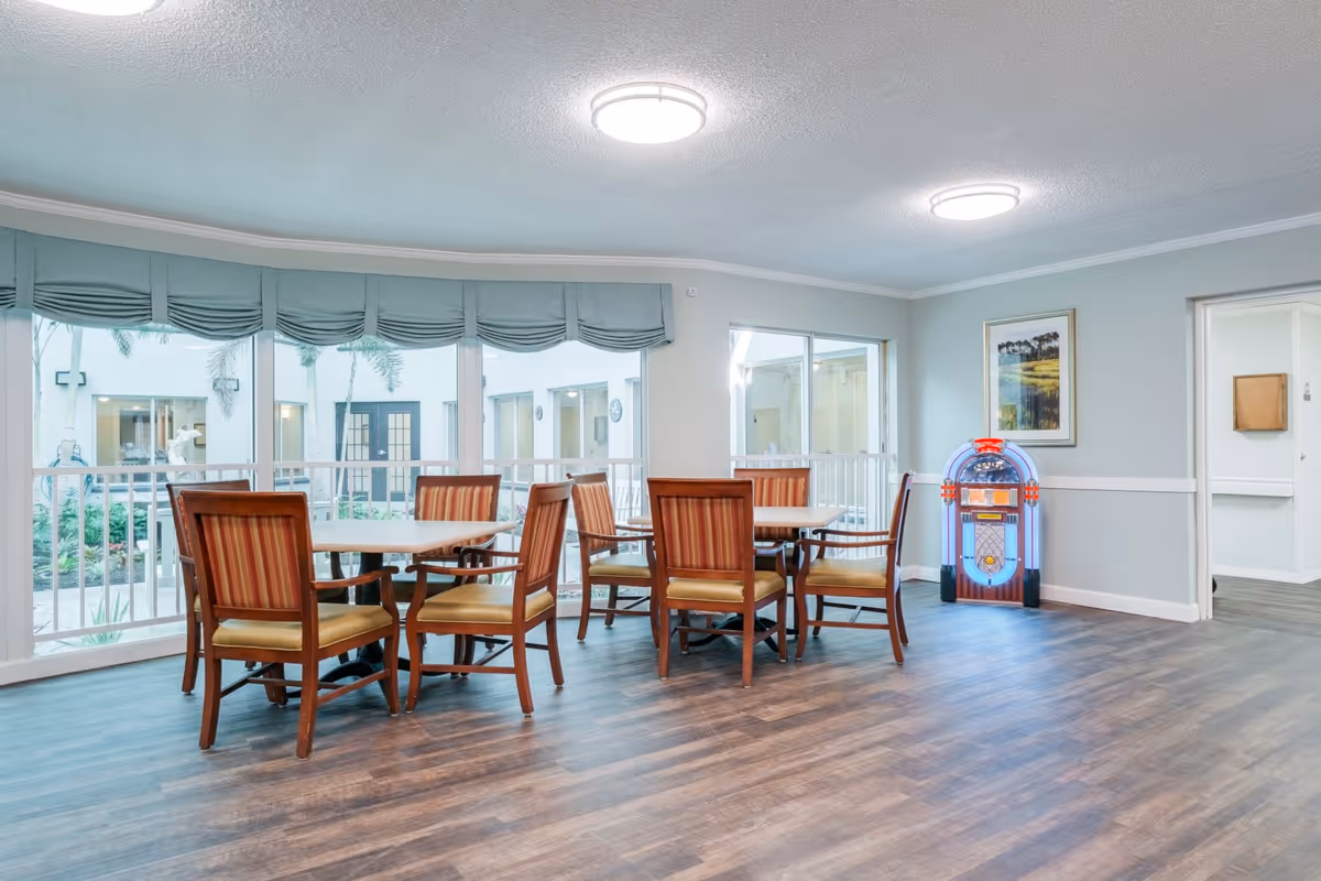 A bright and spacious common area with two wooden tables surrounded by striped cushioned chairs. Large windows with blue valances overlook an outdoor courtyard with palm trees. A vintage-style jukebox with blue and red lights stands against the wall next to a framed landscape painting. The room has wood-look flooring and soft gray walls with white trim.