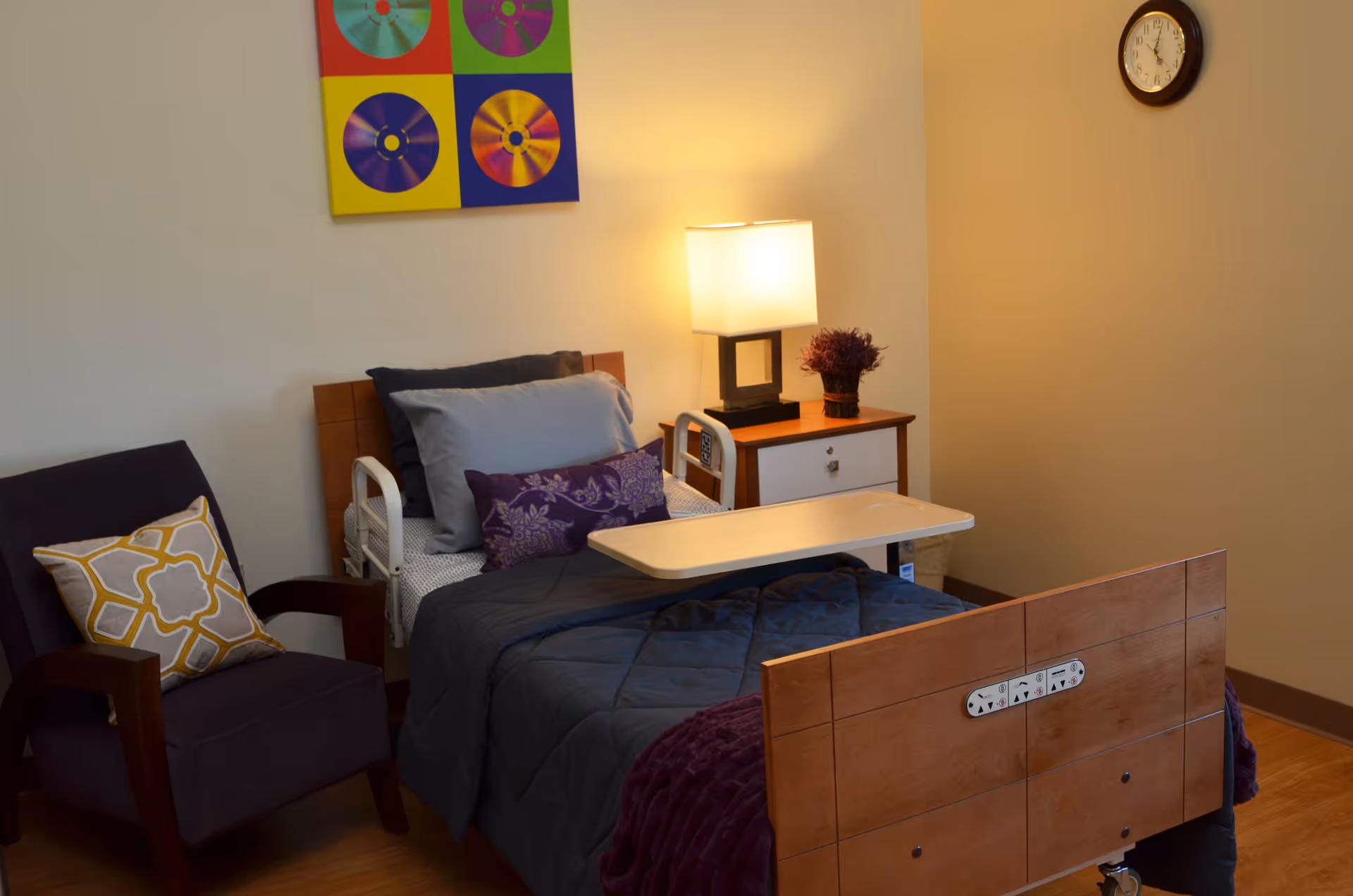 A neatly arranged bedroom in a senior living facility featuring a hospital-style bed with gray and purple bedding, a wooden headboard and footboard, a small overbed table, a wooden nightstand with a lamp and a small plant, a purple armchair with a decorative pillow, a colorful wall art piece, and a clock on the wall.