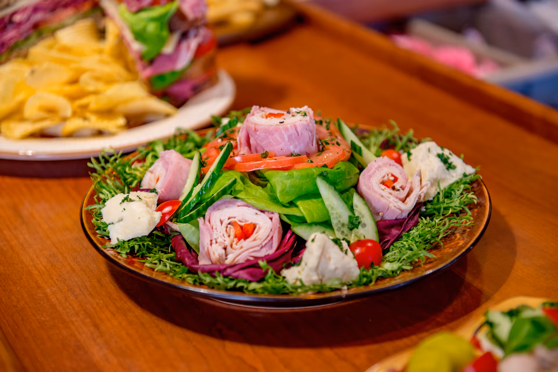 A plate of fresh salad featuring rolled slices of ham, lettuce, tomato slices, cucumber, cherry tomatoes, and dollops of a creamy white dressing, all arranged on a wooden table with another plate of sandwiches and chips in the background.
