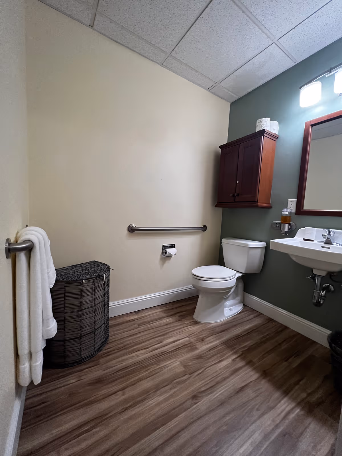 Small accessible bathroom with a toilet, wall-mounted sink and mirror, wooden cabinet, grab bar, towel, and laundry basket on wood-look flooring.