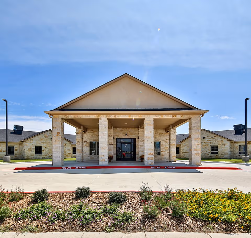 Front exterior view of a single-story building with a covered entrance supported by stone pillars, surrounded by landscaped flower beds and a clear blue sky.