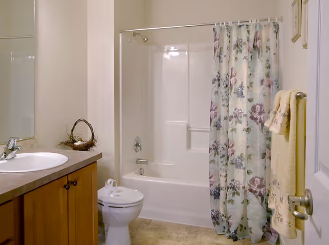 A clean bathroom featuring a white bathtub with a floral shower curtain, a toilet with a decorative item on top, a wooden vanity with a sink, and yellow towels hanging on a towel rack next to the door.