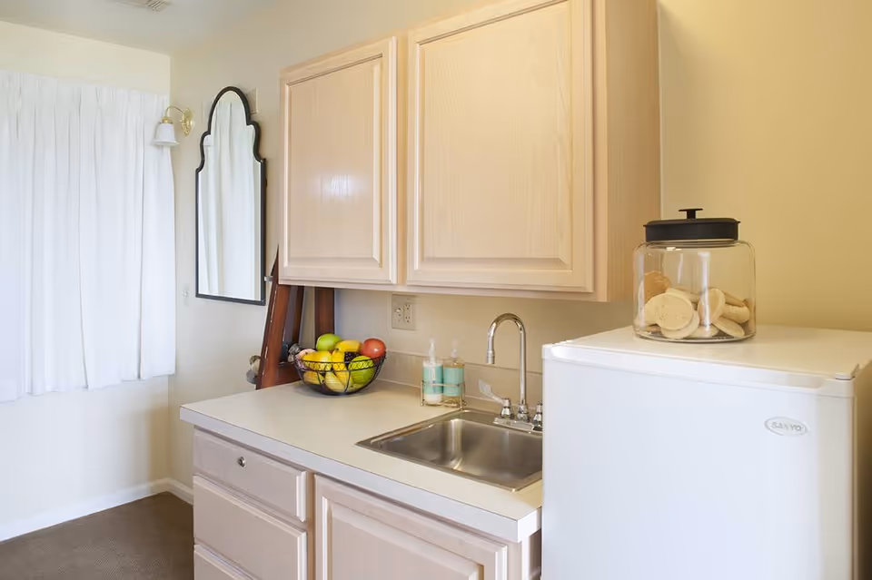 A small kitchenette area with light wood cabinets, a stainless steel sink, a white mini refrigerator with a glass jar of cookies on top, a bowl of assorted fruit on the counter, a wall-mounted mirror with a black frame, and a window with white curtains.