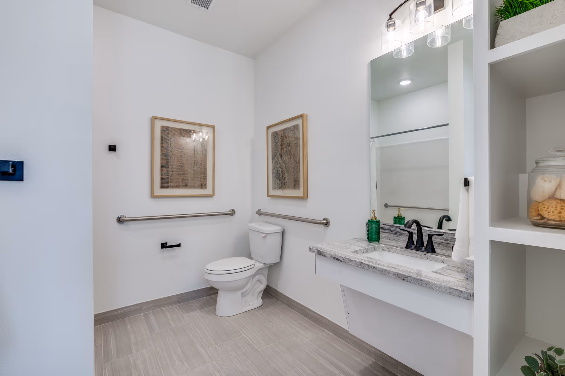 A clean, modern bathroom with a white toilet, two framed artworks on the wall, stainless steel grab bars, a granite countertop with a sink, black faucet, and a large mirror above. There are shelves on the right side with decorative items including a glass jar with sponges and a green plant.