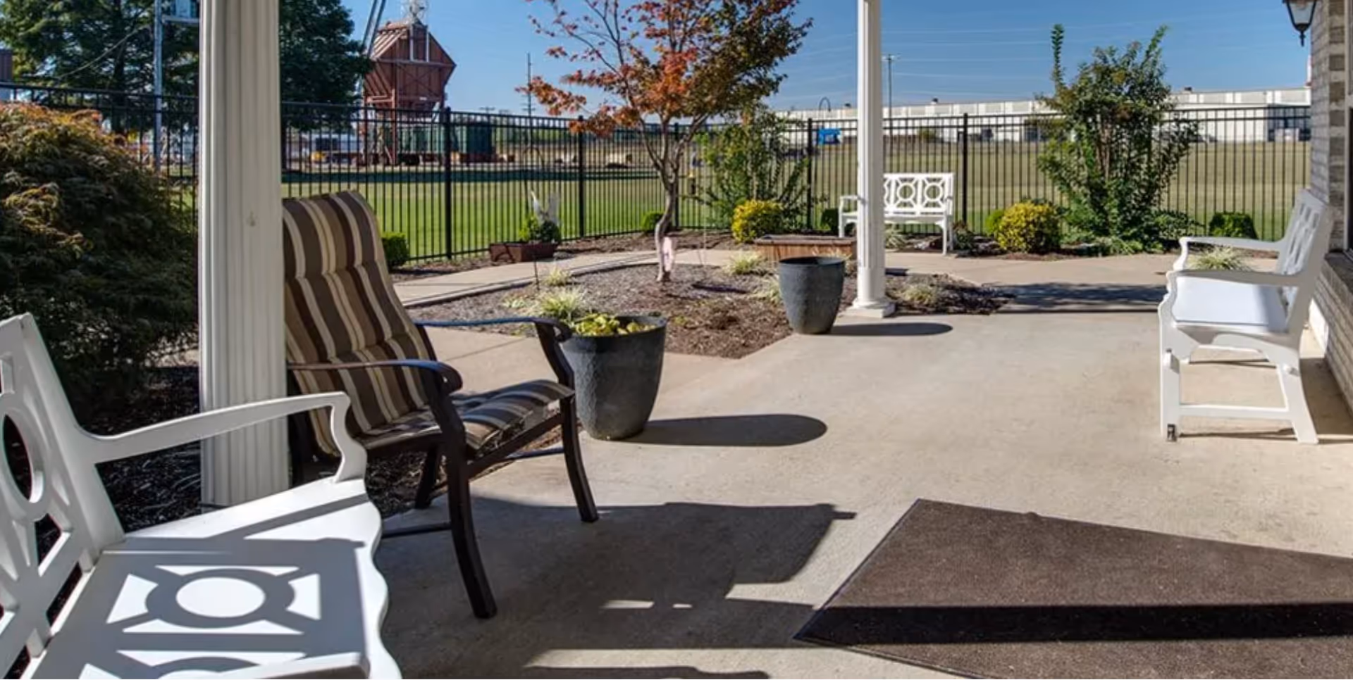 Covered patio with benches, chairs, and potted plants overlooking a fenced lawn and landscaping.