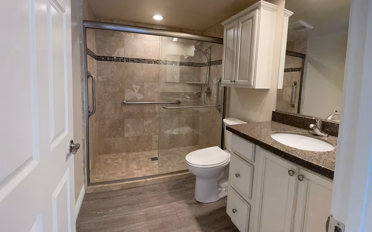 A modern bathroom featuring a glass-enclosed walk-in shower with beige tiles and a decorative horizontal strip, a white toilet, and a vanity with a granite countertop, white cabinets, and a sink. The floor has wood-like tiles, and there is a large mirror above the vanity.