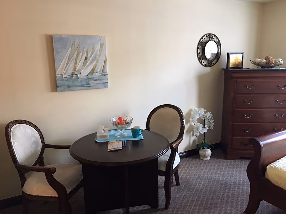 A cozy room with a round wooden table and two upholstered chairs. On the table are a glass bowl with decorative items, a blue mug, and some papers. The room also features a wooden dresser, a bed with a wooden frame, a small potted plant with white flowers, a round decorative mirror on the wall, and a painting of sailboats on the wall.