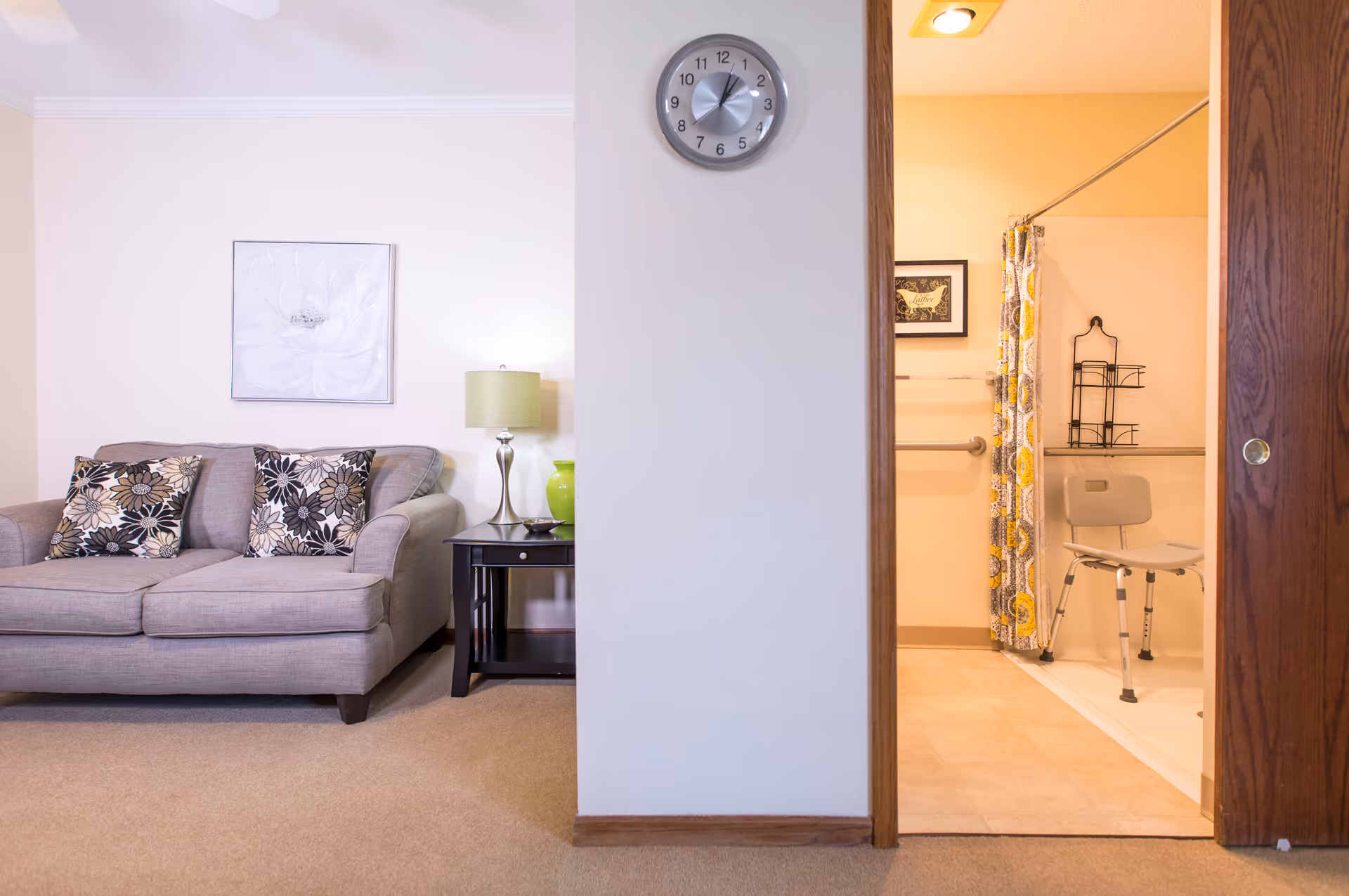 A living room with a gray sofa and side table visible next to an open doorway revealing an accessible bathroom with a shower chair.