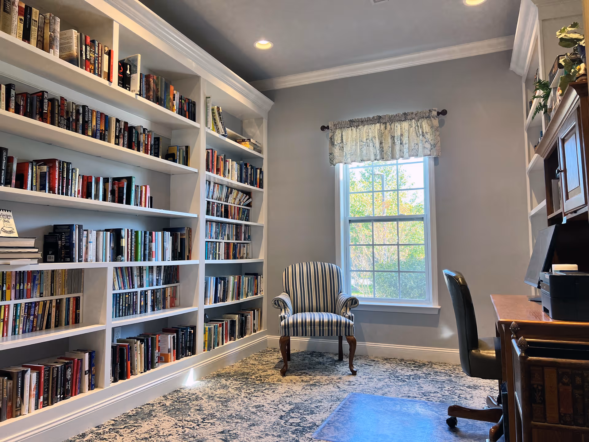 A cozy reading room with built-in white bookshelves filled with books along the left wall, a striped armchair positioned near a window with a floral valance, and a wooden desk with a black office chair on the right side. The room has a patterned carpet and soft natural light coming through the window.