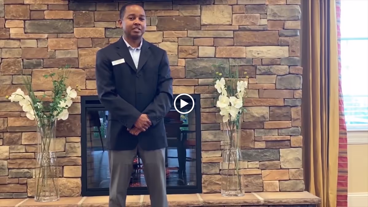 A man wearing a dark blazer and gray pants stands in front of a stone fireplace with two tall glass vases containing white flowers on either side. There is a window with curtains to the right.