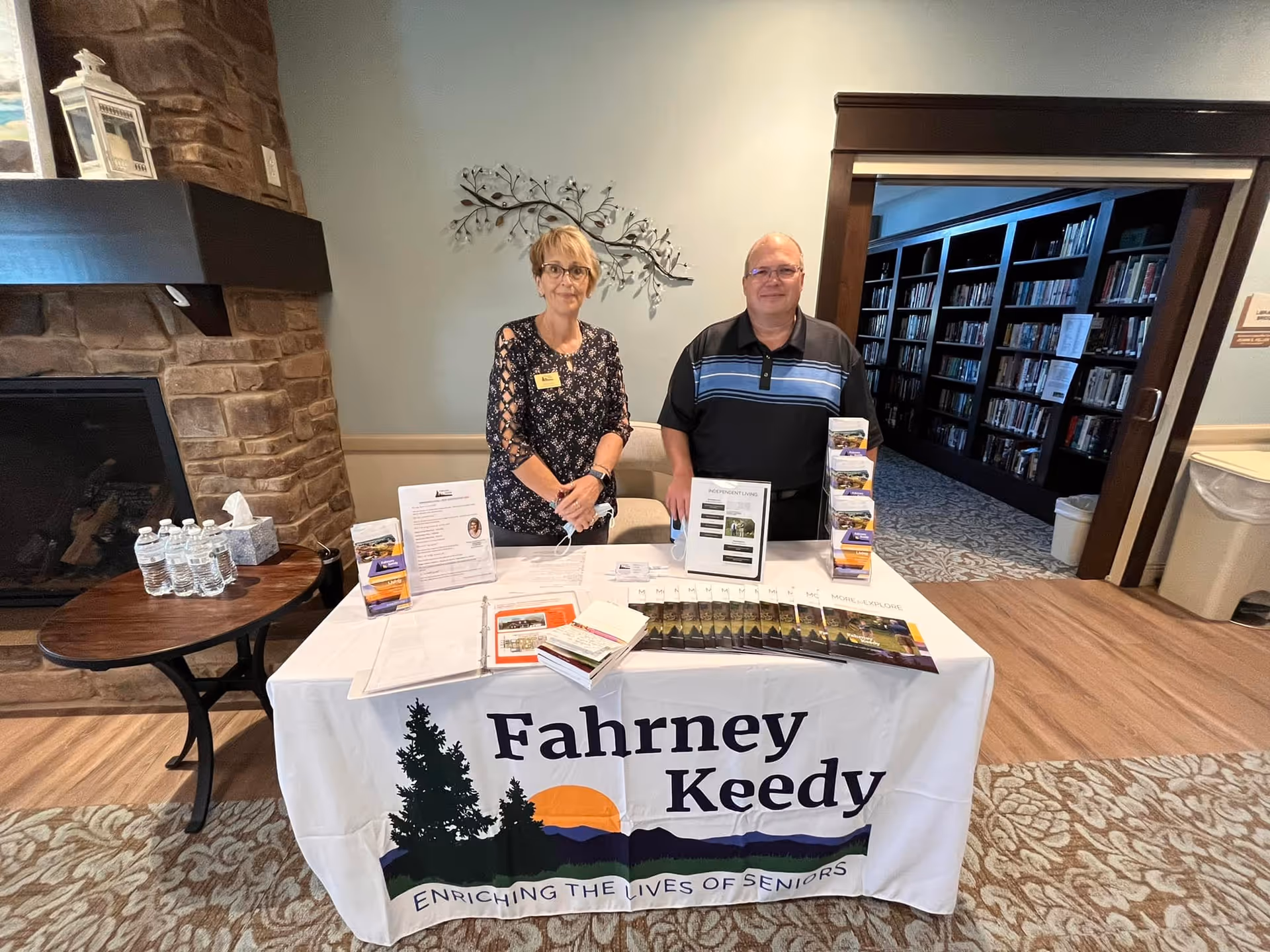 Two people standing behind a table covered with a Fahrney Keedy Senior Living Community tablecloth. The table displays brochures, pamphlets, and informational materials. Behind them is a stone fireplace on the left and a doorway leading to a room with bookshelves on the right.