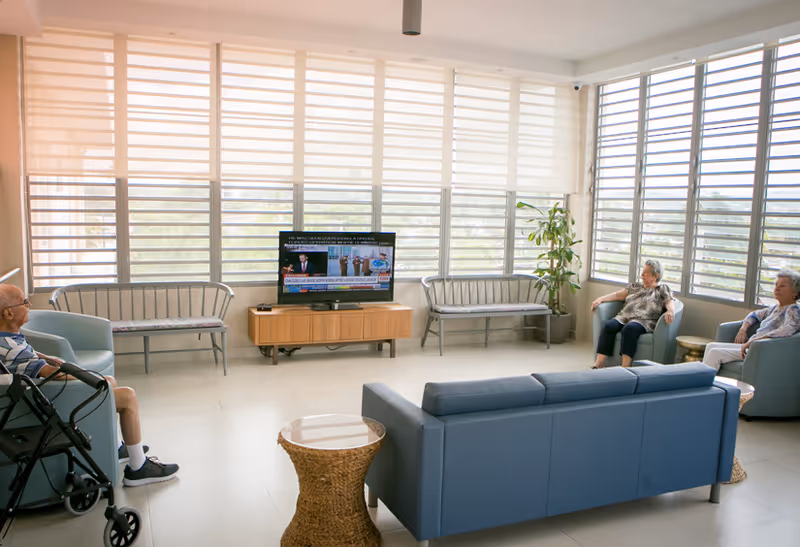A bright and spacious living room in Parkville Home with large windows covered by horizontal blinds. The room has a blue sofa, two blue armchairs, two metal benches, a wooden TV stand with a flat-screen television, a wicker side table, and a potted plant. Three elderly people are seated, watching the television.