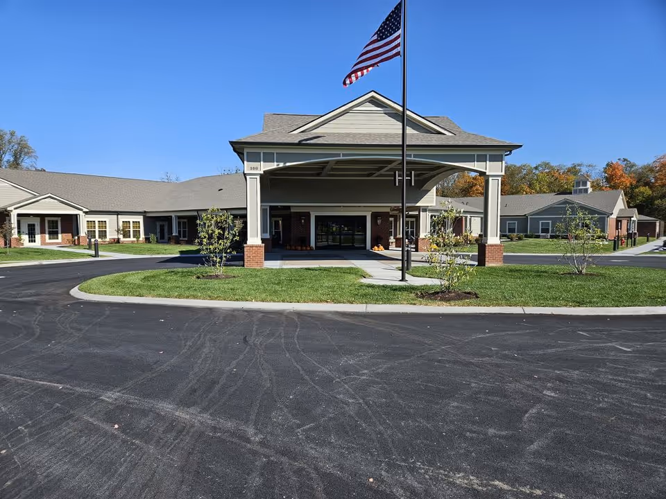 Front entrance of a single-story assisted living building with a covered porte-cochère, American flag, and circular driveway.