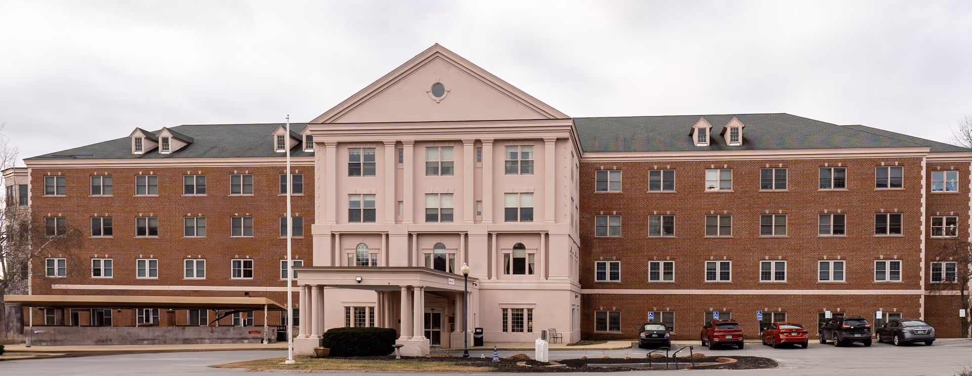Wide brick and pale-pink trimmed multi-story senior living building with a central portico and cars parked in front.