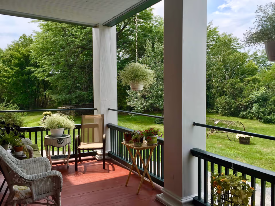 A covered porch with red wooden floor and white pillars overlooking a green garden. The porch has wicker chairs, a wooden chair, a small table with potted plants, and hanging flower pots. Trees and grass are visible in the background under a partly cloudy sky.