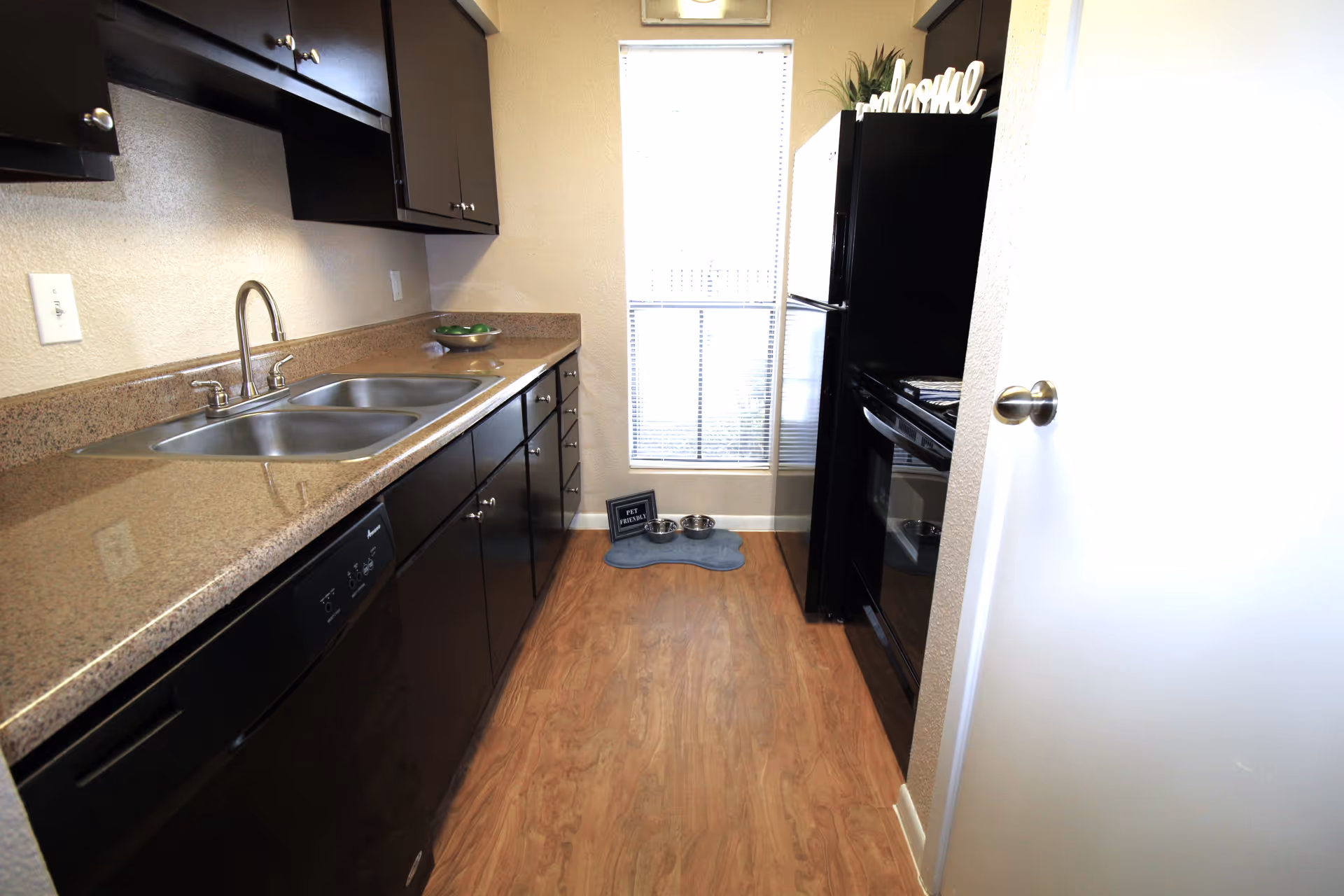 Narrow galley kitchen with dark cabinets, laminate countertops, a double stainless sink, black appliances and wood-look flooring.