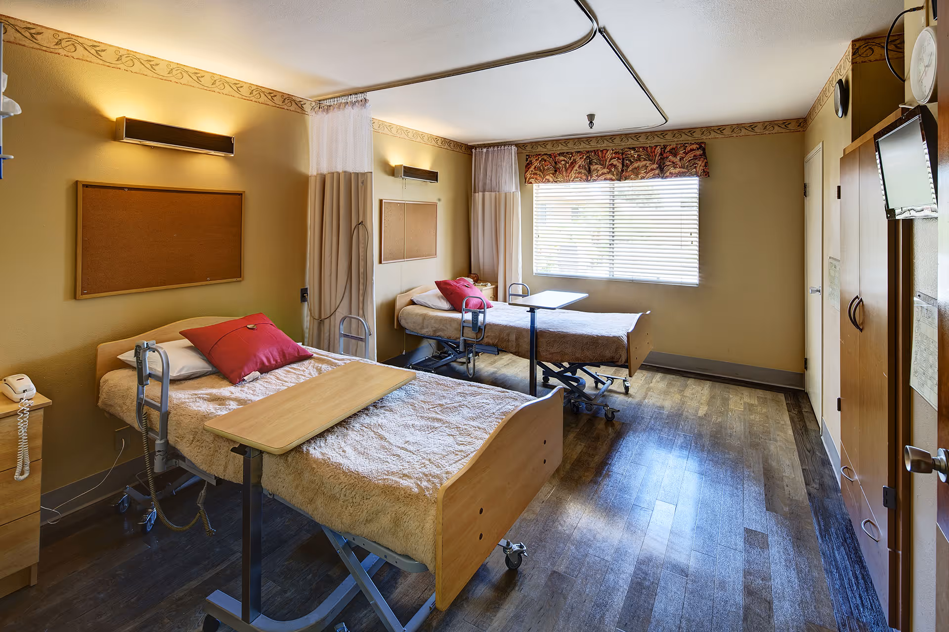 A room with two hospital-style beds, each with a red pillow and beige blanket. Each bed has an adjustable overbed table. The room has wooden flooring, beige walls with a decorative border near the ceiling, and a large window with blinds and a floral valance. There are wall-mounted lights above each bed, a wooden cabinet, a wall-mounted TV, and a telephone on a small bedside table.