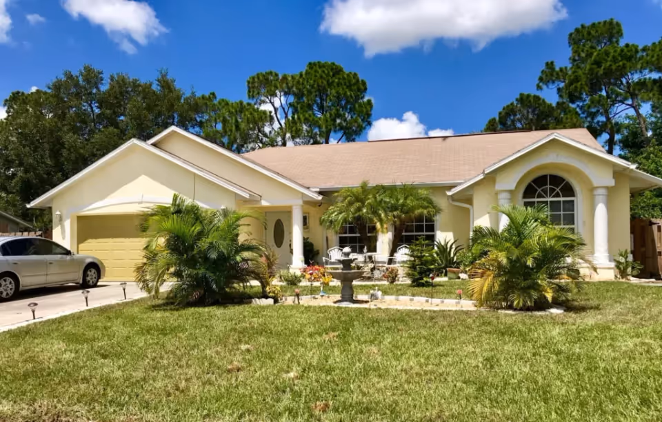Front view of a single-story cream-colored house with a yellow garage, tropical landscaping, and a small decorative fountain in the front yard.