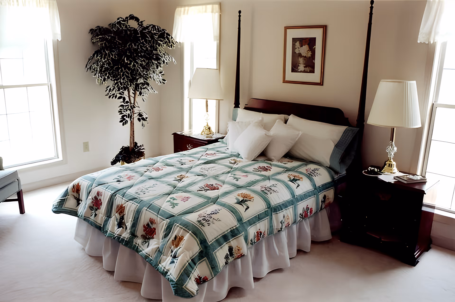 Sunlit bedroom with a four-poster bed dressed in a floral quilt, bedside tables with lamps, and a potted tree by the windows.
