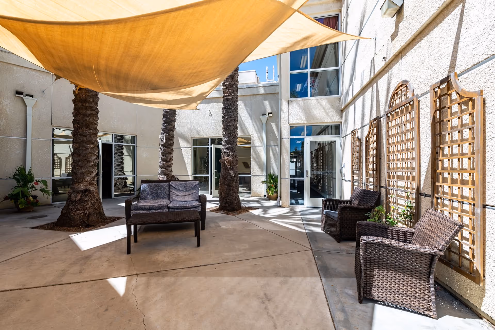 Shaded courtyard with wicker chairs and a loveseat, palm trees, and wooden trellis panels along a building wall.