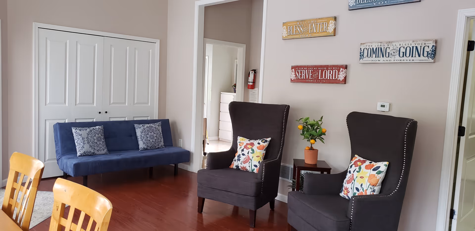 A cozy interior room featuring two dark gray armchairs with colorful floral pillows, a small wooden side table with a potted plant, a blue sofa with two patterned cushions, and wooden dining chairs partially visible in the foreground. The walls are decorated with inspirational wooden signs, and there are white double doors and an open doorway leading to another room.