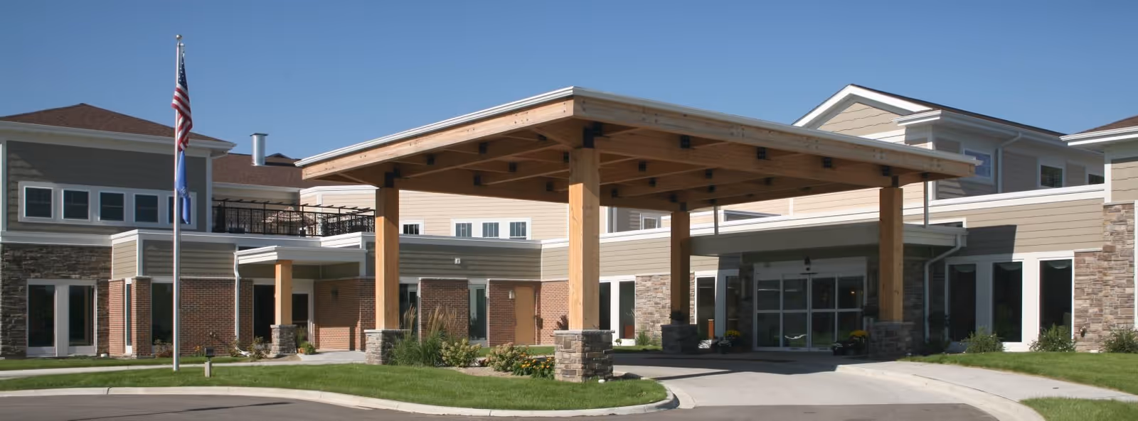 Exterior view of Oak Park Place Janesville senior living facility showing the main entrance with a large wooden canopy supported by stone pillars, surrounded by well-maintained landscaping and an American flag on a flagpole.