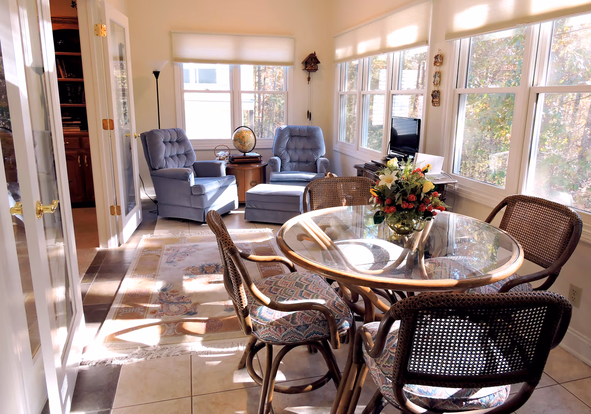 A bright sunlit room with a round glass-top dining table surrounded by four wicker chairs with patterned cushions. In the background, there are two blue cushioned armchairs with an ottoman, a small wooden side table with a globe, a floor lamp, and a TV on a stand. Large windows let in natural light and show trees outside.