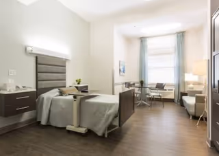A bright, modern nursing facility bedroom with a single bed, bedside tables, a seating area, desk, and a large window with curtains.