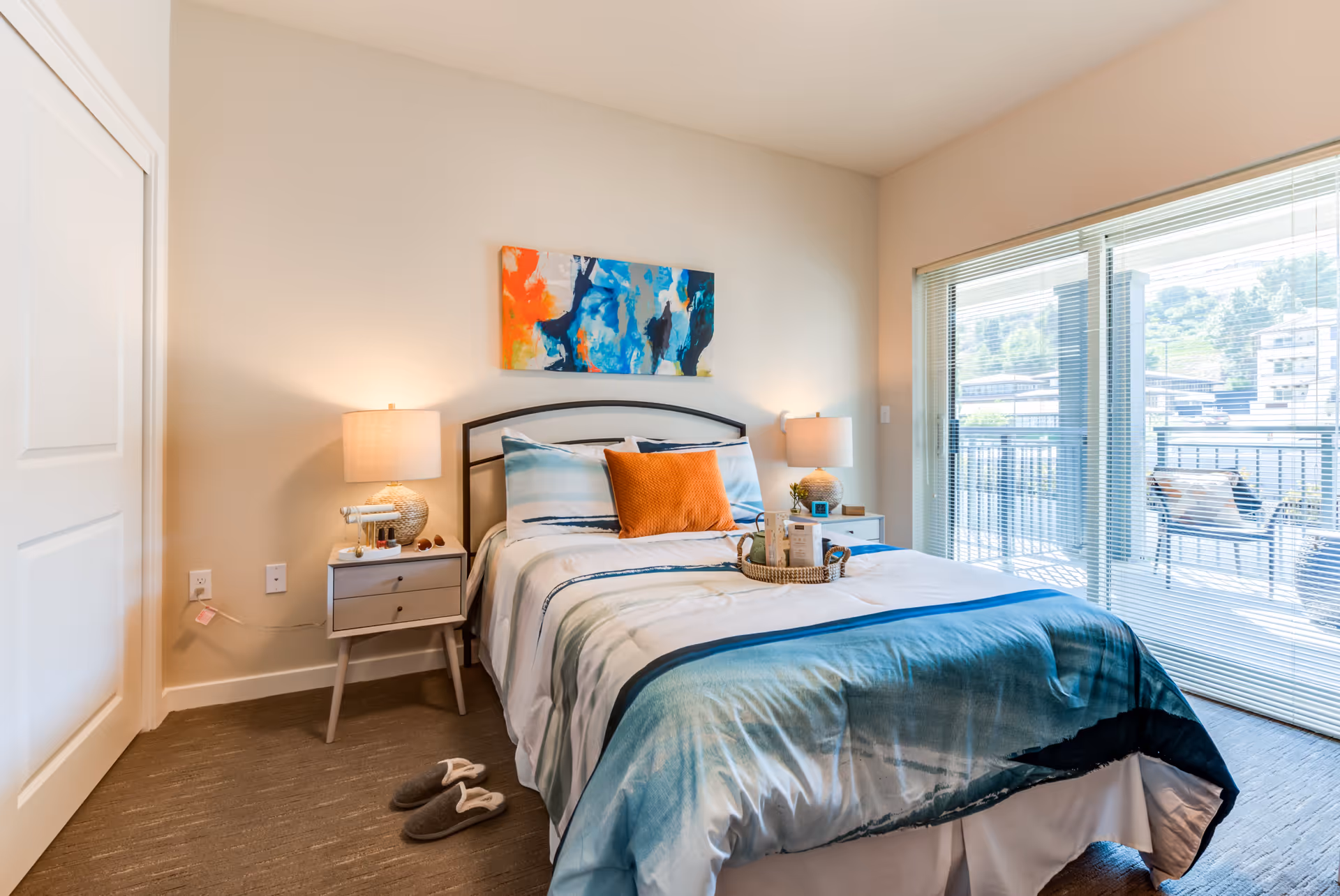 A bright bedroom with a neatly made bed featuring blue and white bedding and an orange accent pillow. Two bedside tables with lamps flank the bed, and a colorful abstract painting hangs above the headboard. Sliding glass doors with blinds lead to an outdoor balcony with chairs and a view of nearby buildings and greenery.