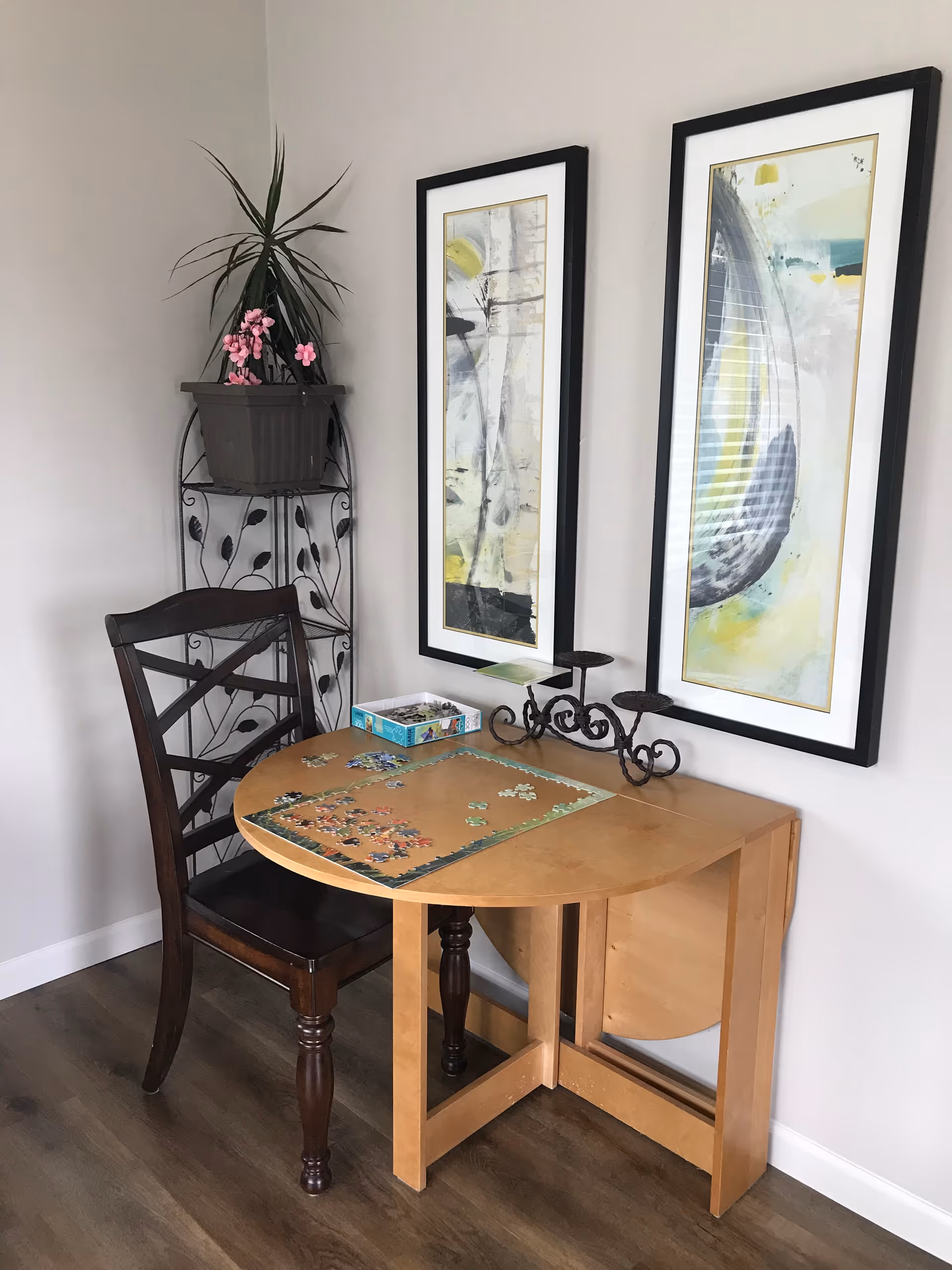 A small wooden table with a partially completed jigsaw puzzle on it, accompanied by a dark wooden chair. Behind the table, there is a decorative metal plant stand holding a potted plant with long leaves and pink flowers. Two framed abstract paintings hang on the light-colored wall above the table.
