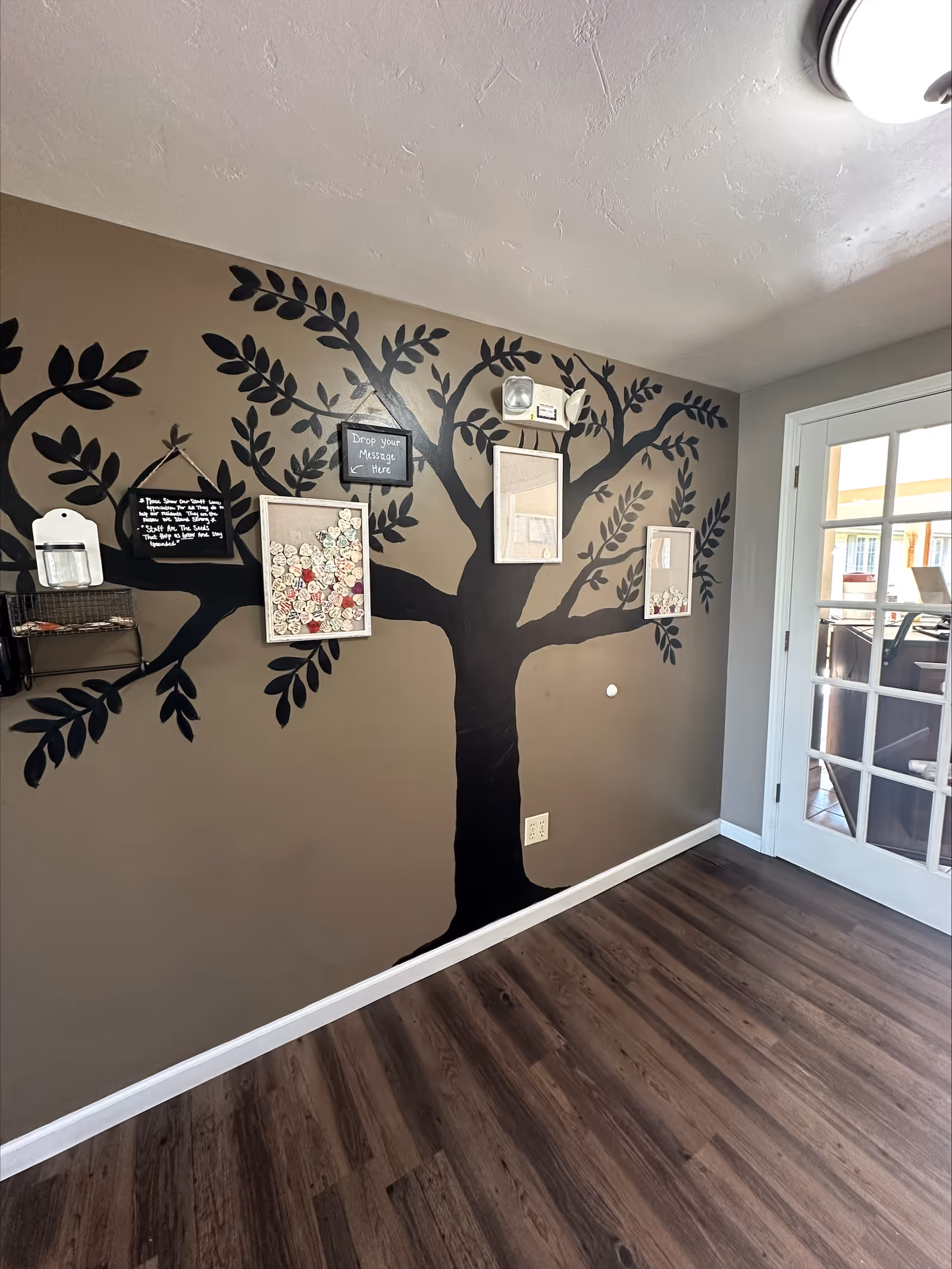 Interior room with a painted black tree mural on a brown wall. The tree has several framed boards attached to it, one with small wooden heart-shaped notes and another with a sign that says 'Drop your Message Here'. The floor is dark wood, and there is a white-framed glass door on the right side leading to another room.