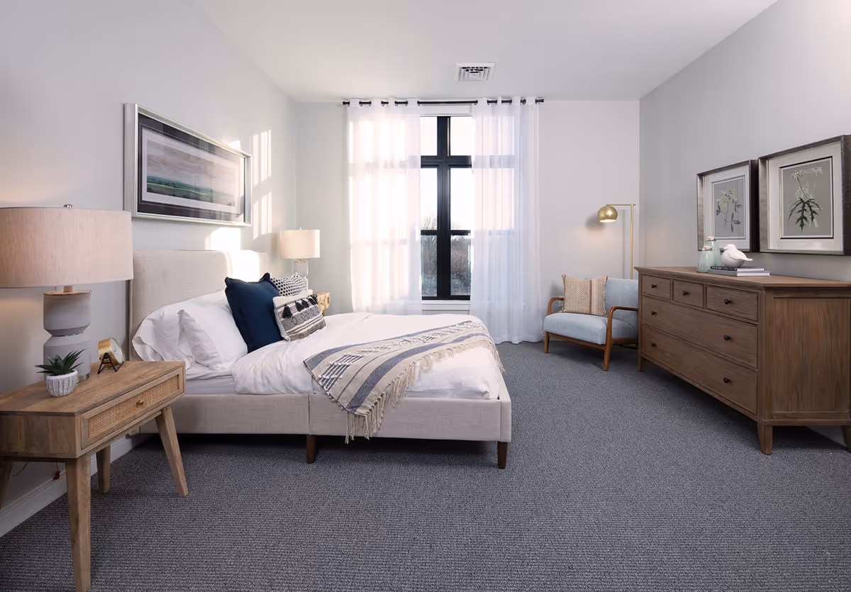 Light-filled furnished bedroom with a bed, nightstand, dresser, armchair, and a large window with sheer curtains.