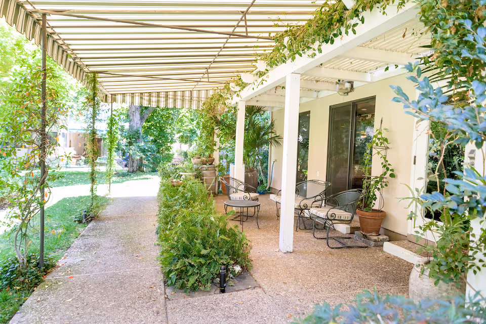 Outdoor patio area with a striped awning overhead, surrounded by lush green plants and trees. The patio has several metal chairs with cushions and small tables, set against the exterior wall of a building with large glass doors and windows.