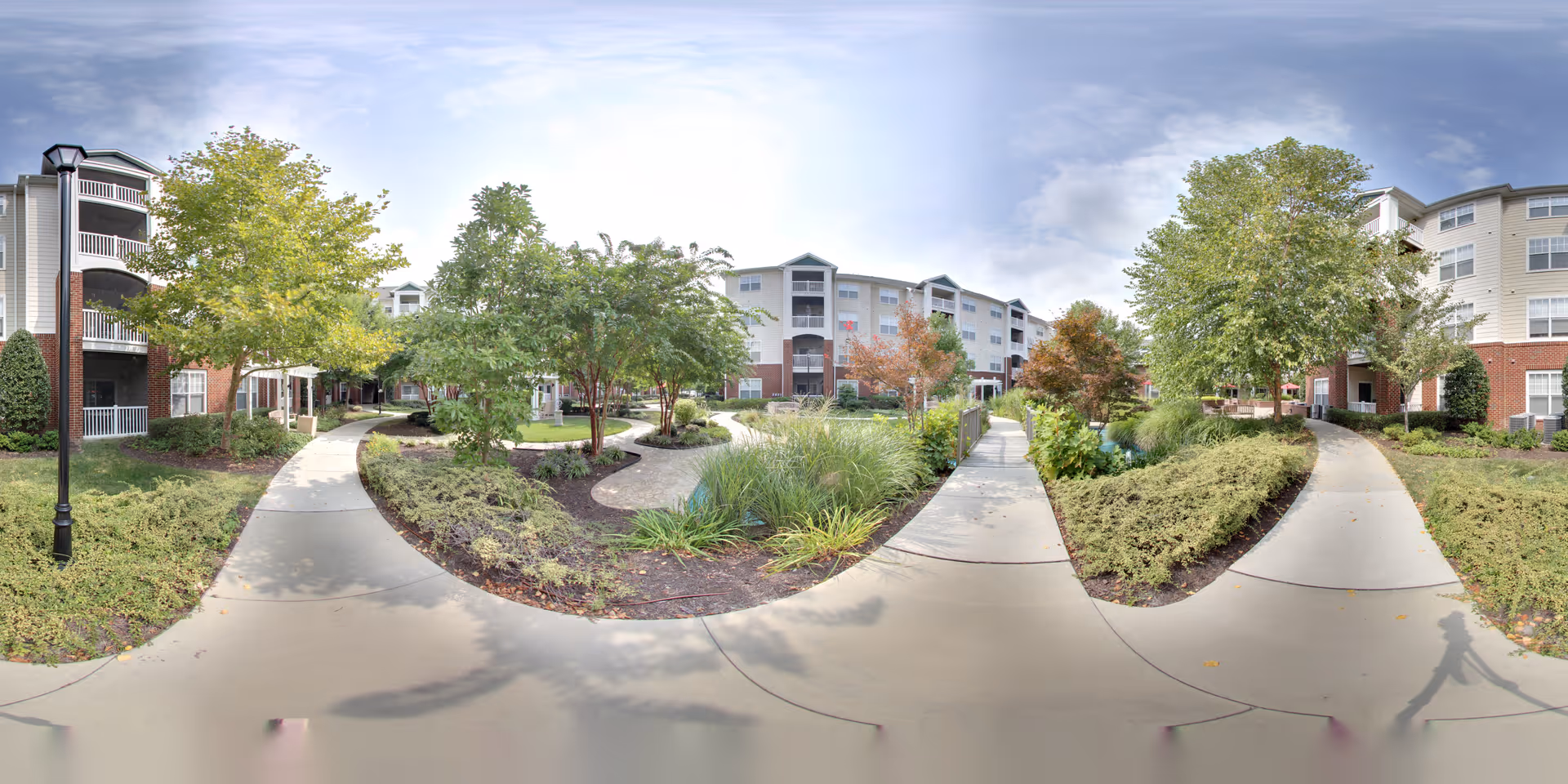 Outdoor view of a senior living facility courtyard with paved walkways, landscaped greenery, trees, and multi-story residential buildings in the background under a partly cloudy sky.