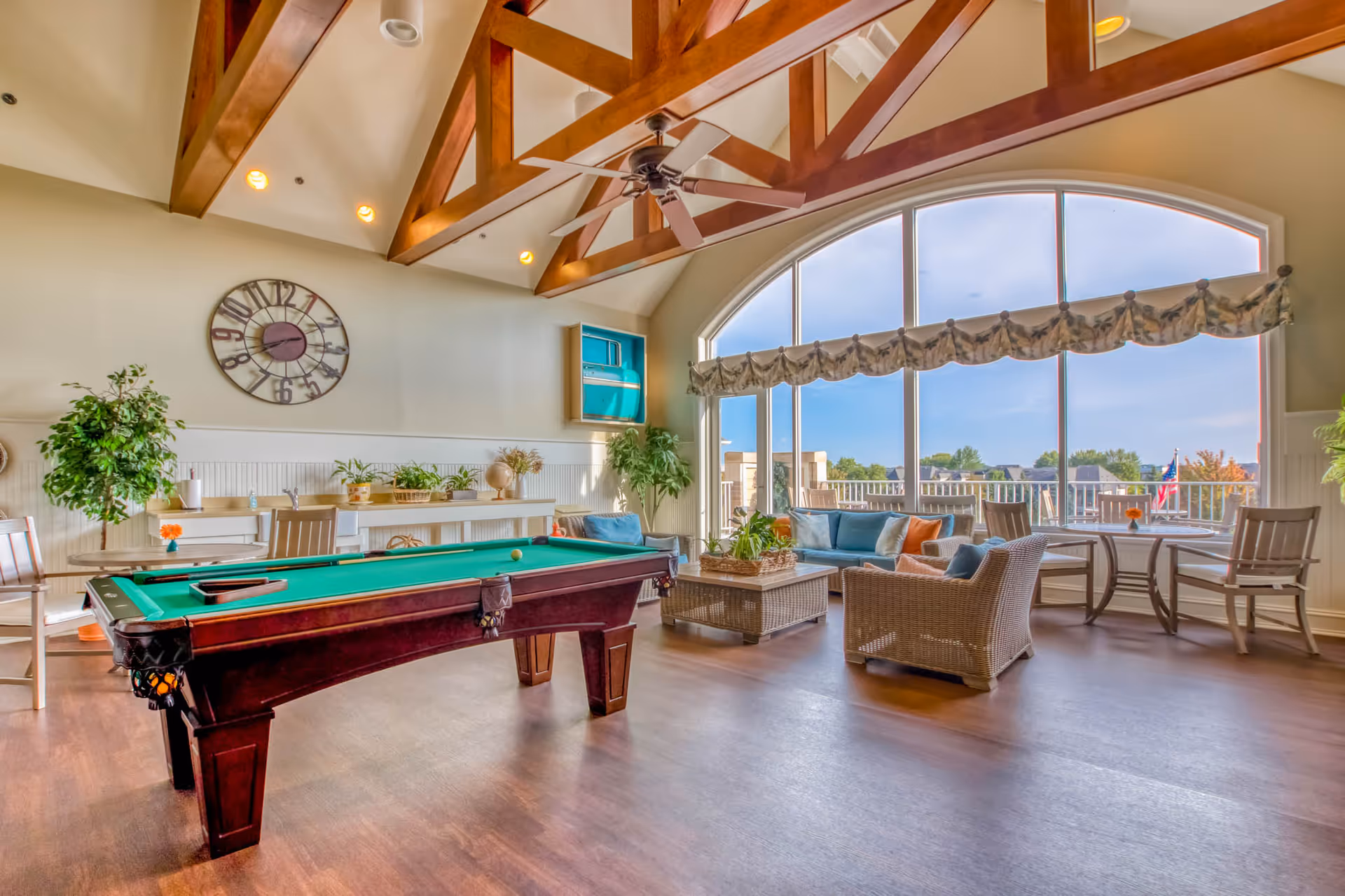 A bright and spacious common area with a pool table in the center, wicker seating with blue cushions near large arched windows, wooden ceiling beams, a ceiling fan, and various plants and decorative items along the walls.