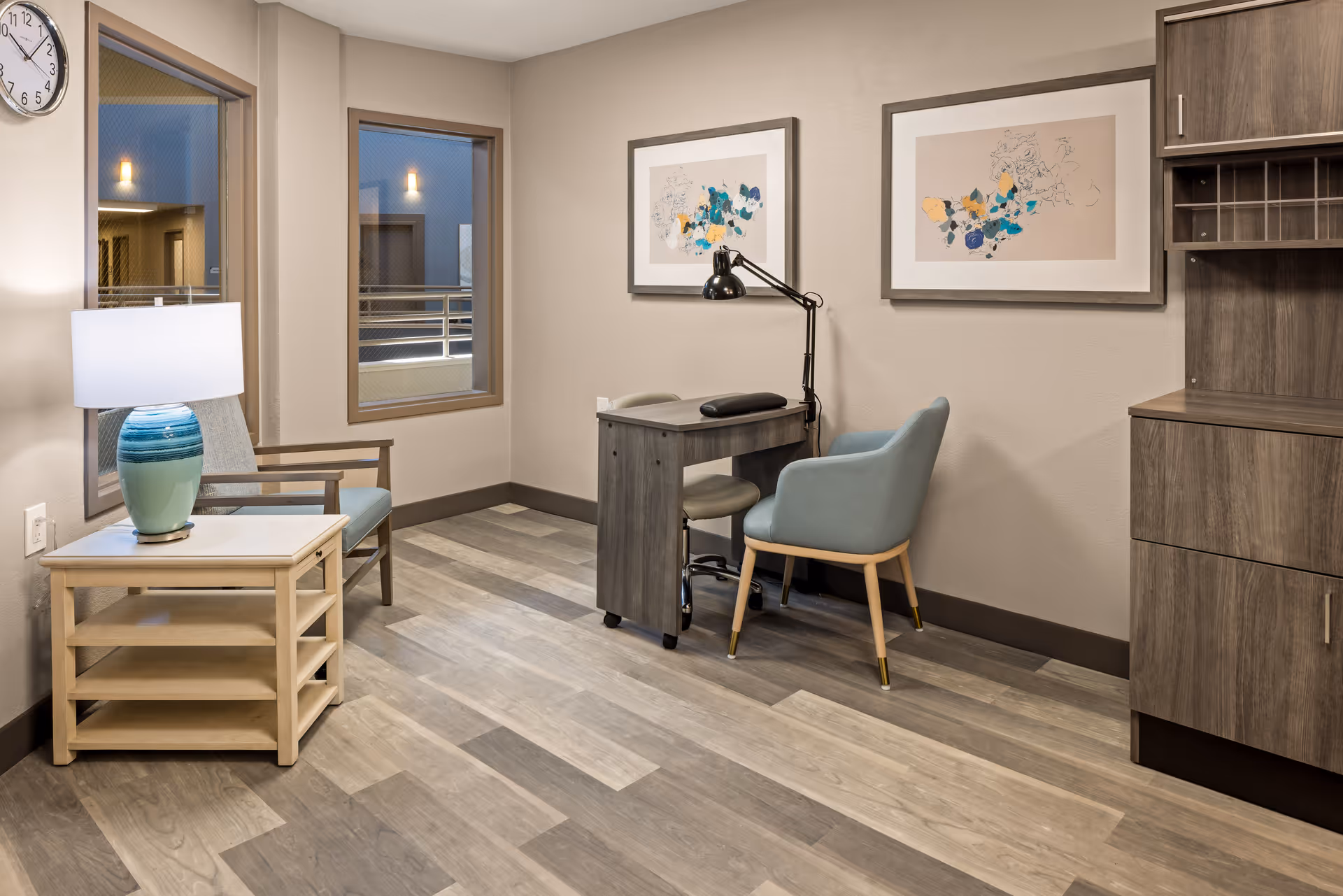 A cozy interior room with wood-patterned flooring, a small wooden desk with a black desk lamp, two chairs including a blue cushioned chair and a wooden armchair, a side table with a blue and white lamp, two framed abstract artworks on the wall, two windows showing an indoor corridor, and a wooden cabinet with shelves and drawers.