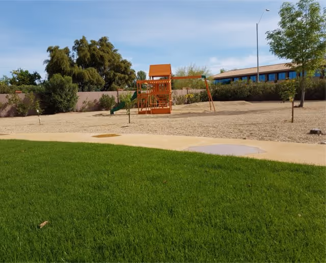 Outdoor courtyard with a small wooden playground, green lawn, walkway, and a building in the background.