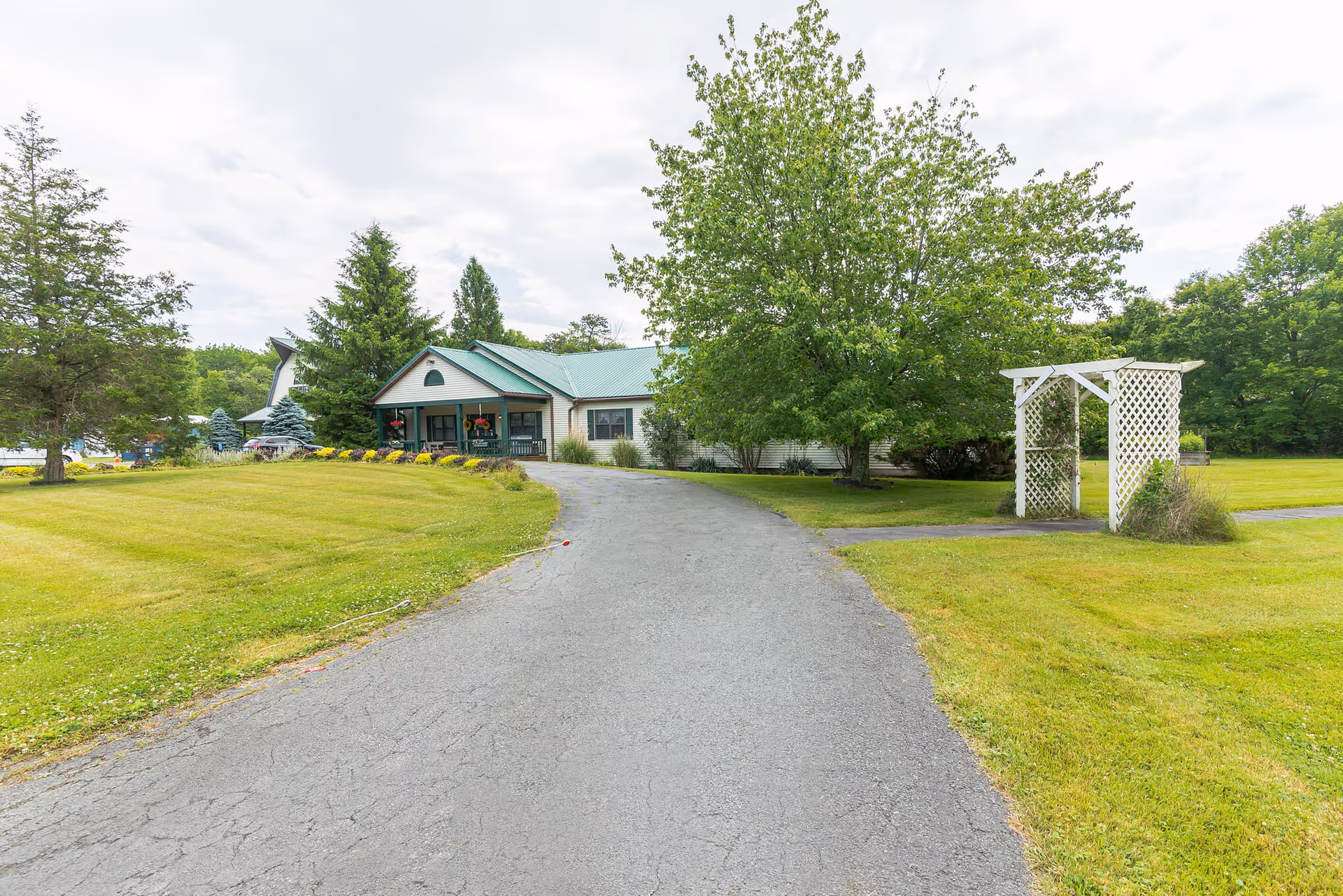 A paved driveway leads to a single-story building with a green roof and a covered porch. The building is surrounded by well-maintained grassy areas, trees, and a white wooden trellis on the right side. The sky is partly cloudy.