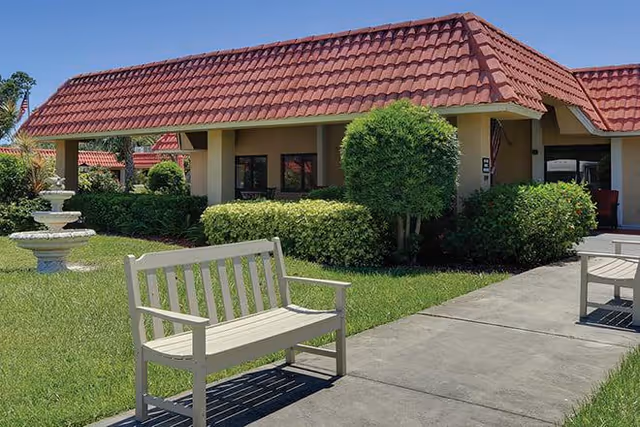Outdoor view of a single-story building with a red tiled roof, surrounded by green bushes and a well-maintained lawn. There is a white bench on a concrete pathway in the foreground and a white tiered fountain on the left side of the image.