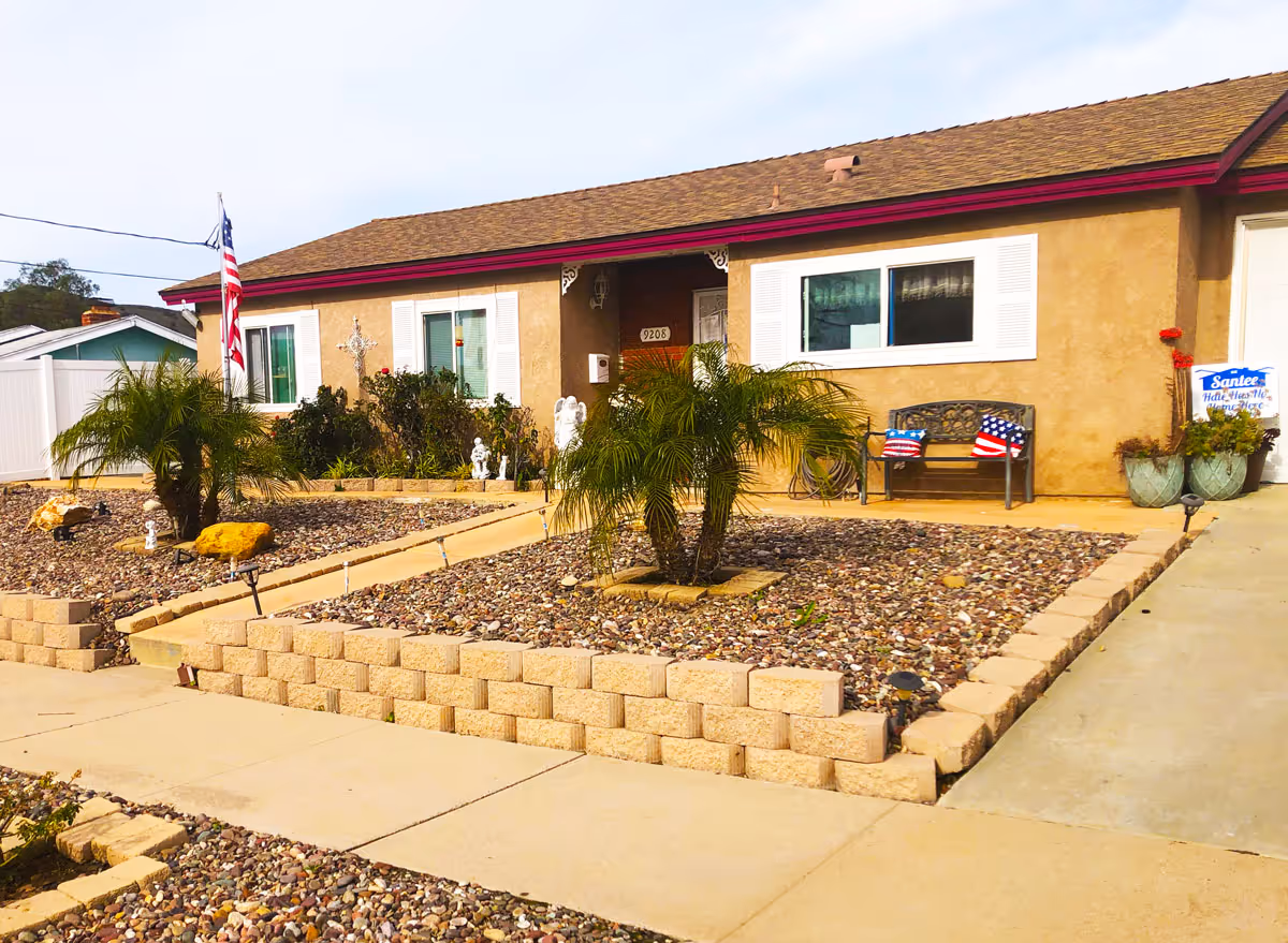 Single-story stucco house front with a low rock garden, small palm trees, American flags, and a bench by the entrance.