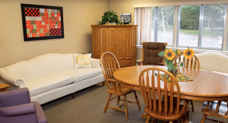 A cozy living room area with a white couch, a purple armchair, a wooden dining table with four chairs, and a wooden cabinet. There is a window with vertical blinds letting in natural light, a framed quilt on the wall, and a vase with sunflowers on the table.