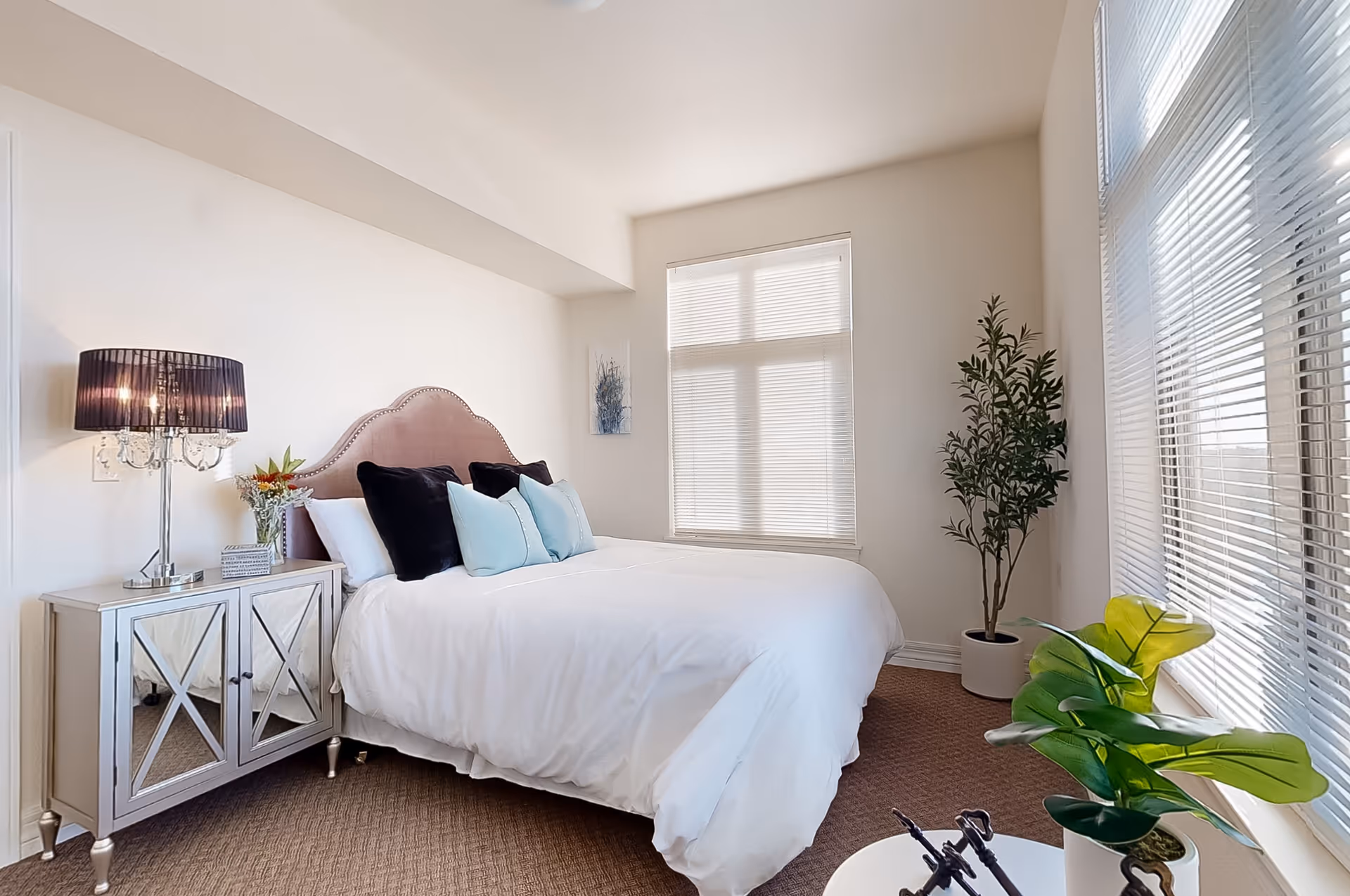 Bright, neatly decorated bedroom with a white bed, mirrored nightstand, lamp, and potted plants by the windows.