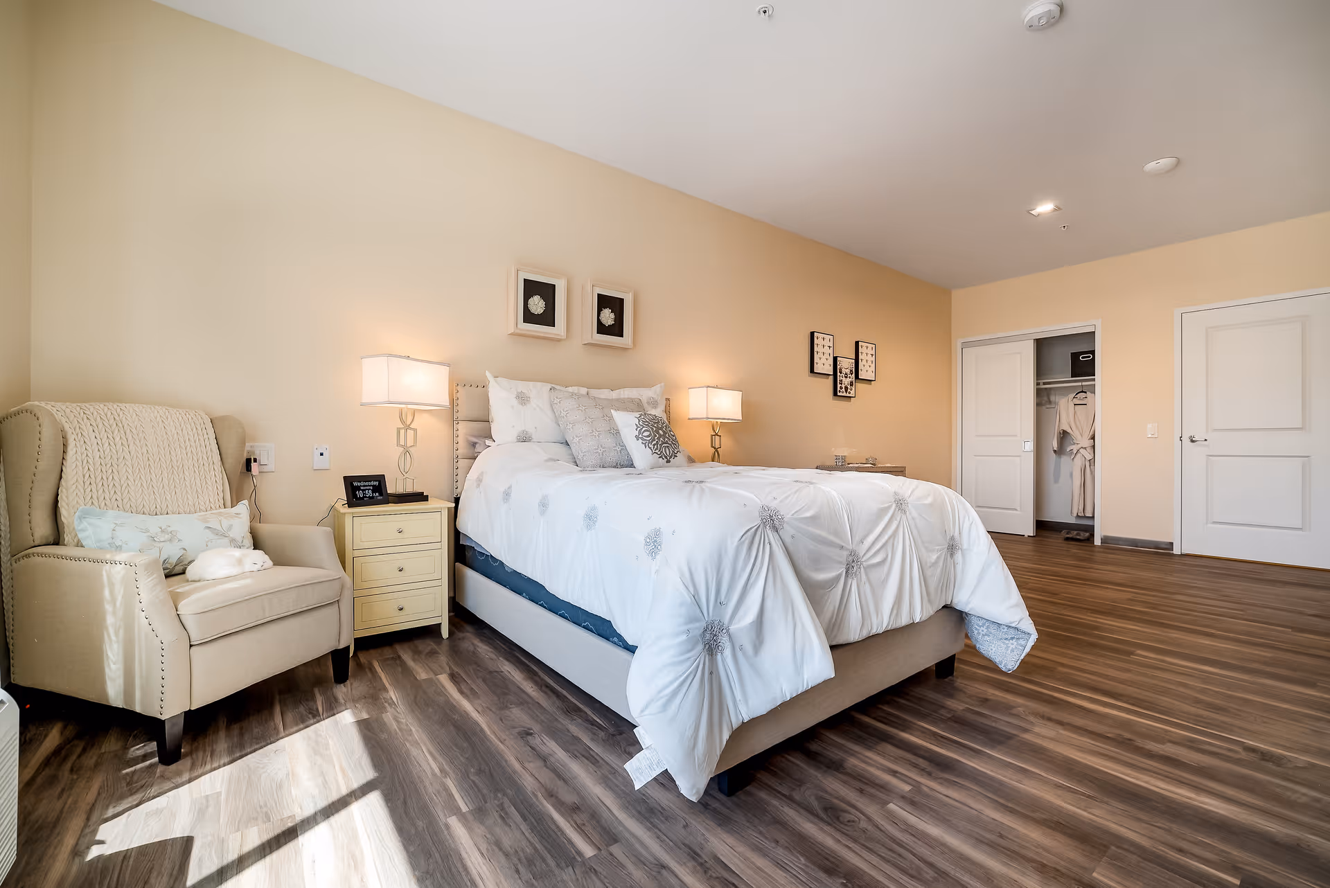 A bright and spacious bedroom with a large bed covered in a white comforter with decorative pillows. There is a beige armchair with a knitted throw and a pillow next to a small yellow nightstand with a lamp and a digital clock. The room has wood flooring, beige walls, framed artwork above the bed, and an open closet with robes hanging inside.