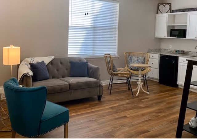 A cozy living area featuring a gray tufted sofa with blue pillows and a throw blanket, a teal upholstered chair, two wicker chairs with a small round white table between them, a floor lamp with a beige shade, and a kitchenette with white cabinets, granite countertop, a microwave, and a mini fridge. The room has wood flooring and a window with white blinds.